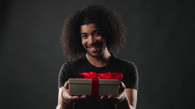 A confident young man with a stylish curly hairstyle and a simple black t shirt smiles genuinely at the camera. The studio portrait features a dark, neutral background, emphasizing the subject s warm 
