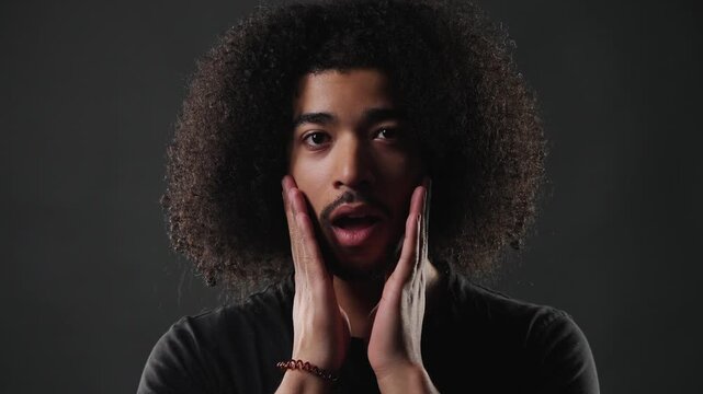 A young man with curly hair expresses surprise and disbelief in a studio setting. His wide eyes and open mouth, coupled with his hands gesturing, convey a strong emotional reaction. This footage is pr