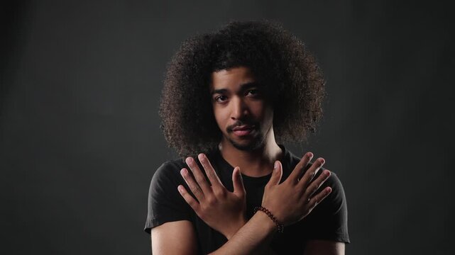 A confident young man with a voluminous afro hairstyle and a dark t shirt poses for a studio portrait. He stands against a simple gray background, looking directly at the camera with a serious and com