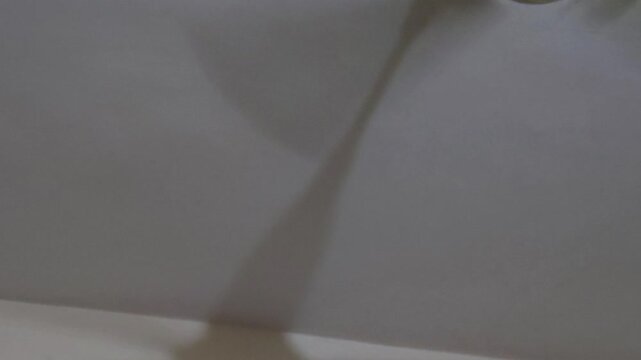 A stark perspective reveals a spinning white ceiling fan casting a distinct shadow on the ceiling. The continuous rotation symbolizes the worsening global climate crisis and unbearable indoor heat wav