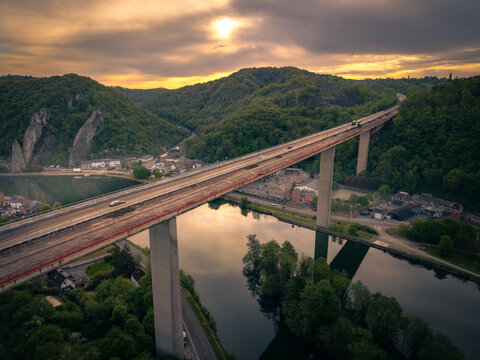 Aerial view of the Charlemagne Viaduct crossing the Meuse river valley surrounded by lush green hills under a golden sunset sky in Dinant, Wallonia, Belgium.