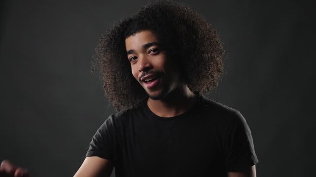 A young man with voluminous curly hair smiles genuinely while looking directly at the camera, conveying youthful confidence and positive emotion. This studio headshot features a dark background, empha