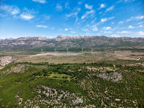 Aerial view of the Suncana elektrana Obrovac solar farm and wind turbines nestled in the rugged karst landscape under a bright blue sky in Zaton Obrovacki, Zadar County, Croatia.