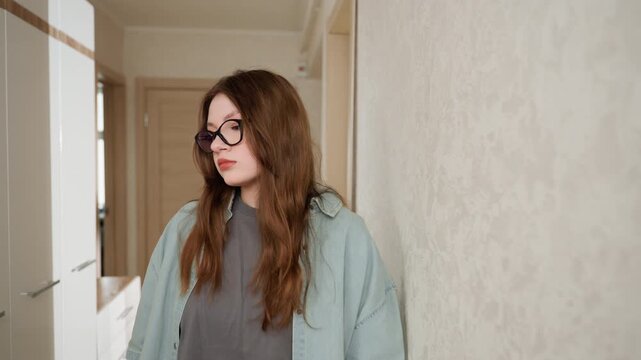 Young caucasian woman standing in hallway, wearing glasses and layered jacket, leaning against textured wall, subdued lighting, introspective expression, casual trousers, home corridor setting, soft