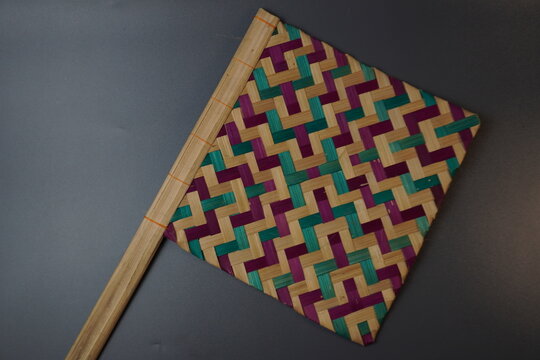 Traditional Indonesian woven bamboo hand fan isolated on a grey background. Colorful satay fan or kipas sate made from braided bamboo skin with geometric pattern, authentic cultural handicraft.