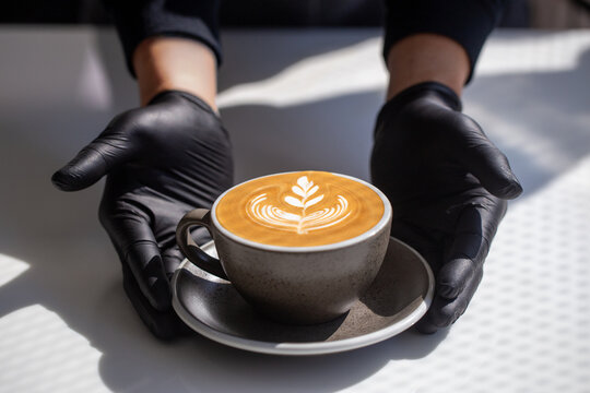 Barista hands in black gloves serving hot latte art coffee cup on white table