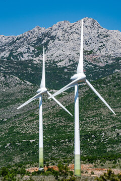 Aerial view of modern wind turbines standing against the rugged rocky peaks of the Velebit mountains under a clear blue sky in Zaton Obrovacki, Zadar County, Croatia.