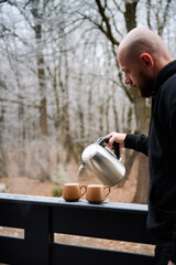 Obraz premium Adult male preparing warm drink with kettle on balcony surrounded by trees, symbolizing relaxation, slow living, weekend retreat, and peaceful countryside moments