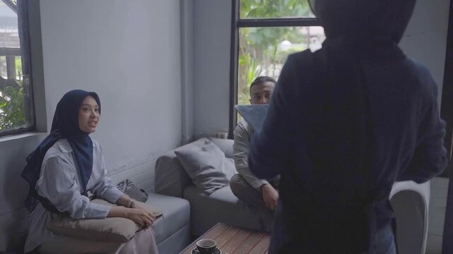 Asian couple seated by window chatting, woman wearing hijab smiles as male guest arrives in casual modern interior with natural