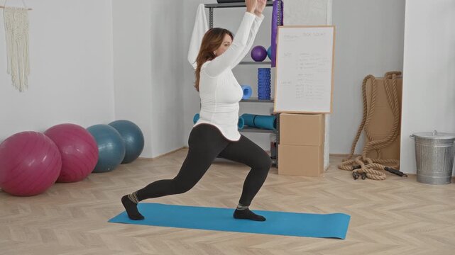 Woman lunges on blue mat in studio with arms raised overhead, transitioning to palms together in a prayer pose beside exercise balls and ropes; focus balance serenity.
