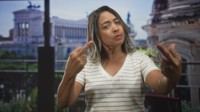 Woman shows both middle fingers to camera, hands raised, wearing a striped shirt on a balcony in front of a historic building facade; defiance rebellion.