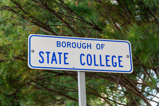 Borough of State College road sign featuring bold blue text on a reflective white surface, clearly identifying the central Pennsylvania community amidst lush green pine tree foliage.