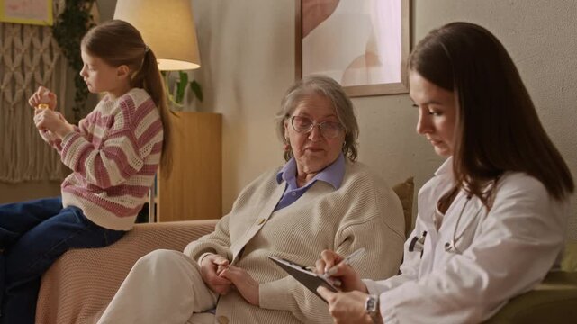 Caucasian doctor taking notes while communicating with senior woman. Little girl opening bottle of pills in background