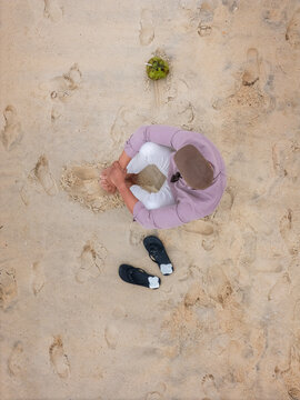 Aerial view of a man sitting on a sandy beach next to a green coconut and flip-flops with footprints in the sand Rio de Janeiro, State of Rio de Janeiro, Brazil.
