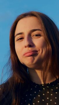 Playful woman making faces with cone outdoor golden hour scene, caucasian model sticks tongue and pouts while holding waffle cone, polka dot blouse, cheek dimples, candid fashion vibe, warm light