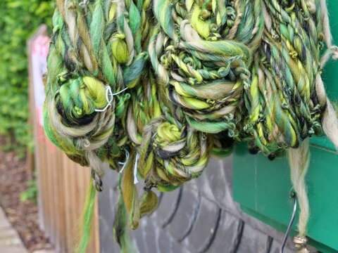 A close-up shot captures thick, textured bundles of hand-spun art yarn hanging
outdoors. The chunky skeins feature a rustic blend of olive green, emerald, lime,
beige, and black threads with uneven sl