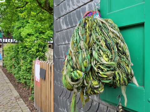 A close-up shot captures thick, textured bundles of hand-spun art yarn hanging
outdoors. The chunky skeins feature a rustic blend of olive green, emerald, lime,
beige, and black threads with uneven sl