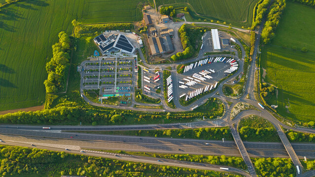 Aerial view of Rugby Services motorway station with solar panels, car parks, and rows of trucks surrounded by green fields and the M6 motorway in Rugby, England, United Kingdom.