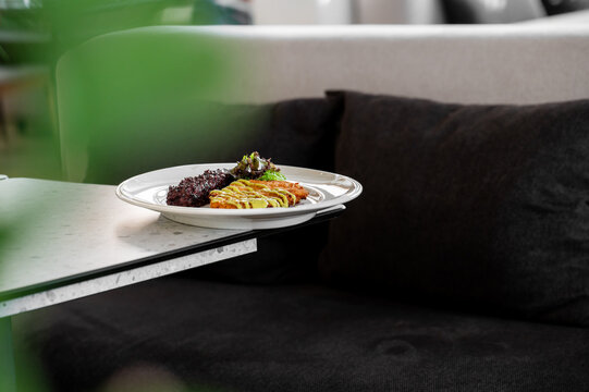 A plate with breaded chicken cutlet drizzled with yellow sauce, dark rice, and fresh greens on a modern table next to a dark sofa.