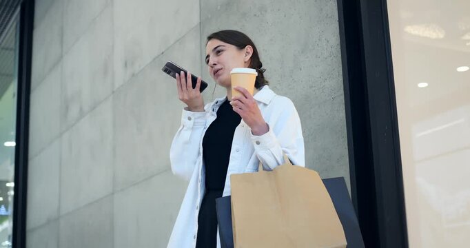 Woman orders coffee. Woman with coffee and shopping bag. Woman on phone holding coffee next to store window. Customer using phone while holding coffee and shopping bag outside storefront