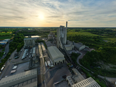Aerial view of the Rugby Cement Plant industrial complex with its tall chimney and silos surrounded by green fields at sunset in Rugby, England, United Kingdom.