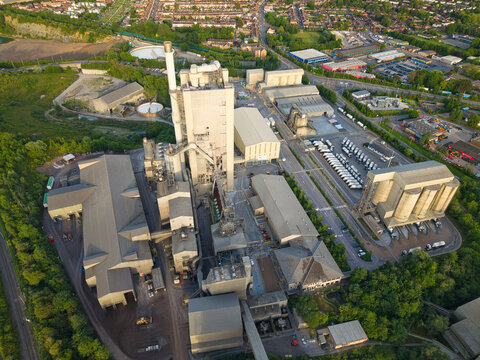 Aerial view of the Rugby Cement Plant featuring industrial buildings, a tall chimney, and silos surrounded by lush greenery and residential areas in Rugby, England, United Kingdom.