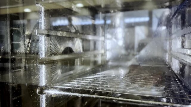 Inside view of a commercial combi oven washing cycle in a restaurant kitchen. Professional steamer oven self-cleaning mode with high-pressure water spraying on stainless steel racks and glass door