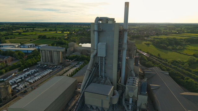 Aerial view of the Rugby Cement Plant with tall chimneys and silos surrounded by green fields during a golden sunset Rugby, England, United Kingdom.