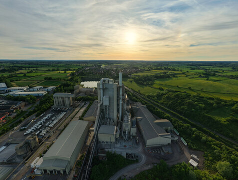 Aerial view of the Rugby Cement Plant with industrial structures and tall chimneys surrounded by lush green fields during a golden sunset Rugby, England, United Kingdom.