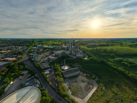 Aerial view of the Rugby Cement Plant industrial complex with its tall chimney tower surrounded by green fields and roads under a golden sunset sky in Rugby, England, United Kingdom.