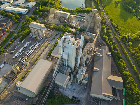 Aerial view of the Rugby Cement Plant featuring its massive industrial towers and silos surrounded by lush green fields and a small lake in Rugby, England, United Kingdom.