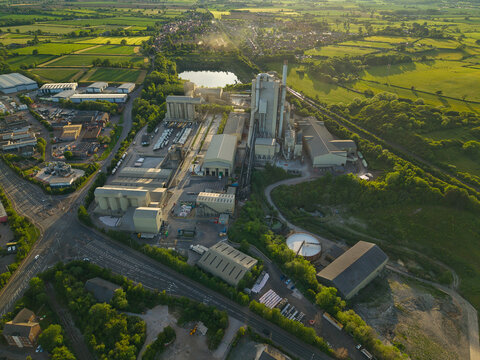 Aerial view of the Rugby Cement Plant featuring industrial silos and tall chimneys surrounded by lush green fields and a small lake at sunset Rugby, England, United Kingdom.