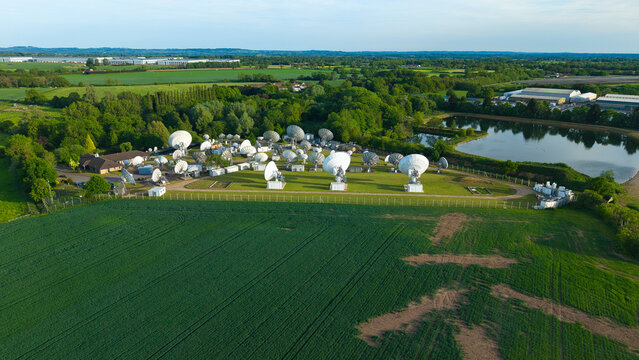 Aerial view of the Satellite Mediaport featuring numerous large white parabolic antennas surrounded by green fields and a small pond Long Lawford, England, United Kingdom.