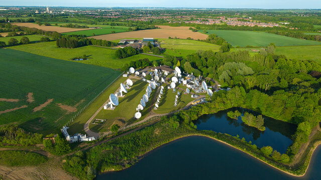 Aerial view of Satellite Mediaport with numerous large white satellite dishes surrounded by lush green fields, dense trees, and a calm pond Long Lawford, England, United Kingdom.