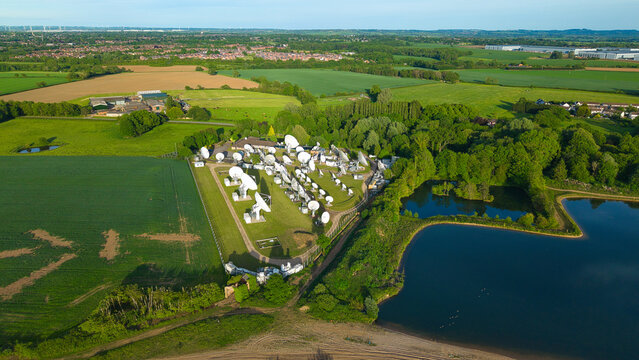 Aerial view of the Satellite Mediaport featuring numerous white parabolic antennas surrounded by lush green fields and a small lake in Long Lawford, England, United Kingdom.