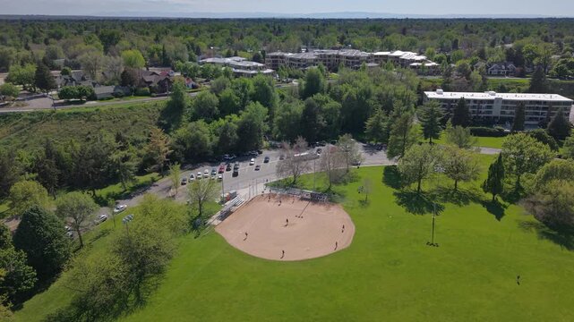 Slow pull back drone shot of softball players actively playing in the ball diamond at Ann Morrison Park, Boise, Idaho. Dynamic game action on a sunny spring day.