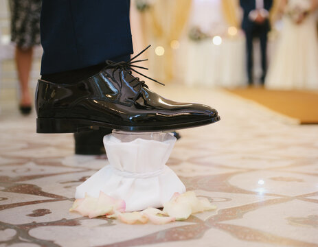 Groom's foot in a polished black shoe stepping on a glass wrapped in a white cloth during a traditional ceremony.