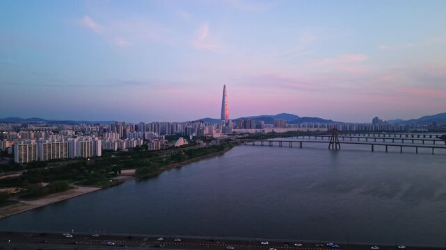Aerial pullback revealing traffic on Cheonho Bridge with pink sunset reflections glowing on Lotte World Tower over the Han River in Seoul, South Korea