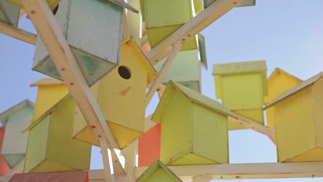Many vibrant wooden birdhouses in various pastel colors are arranged together on a structure against a bright blue sky, creating a whimsical outdoor art installation focused on nature and home
