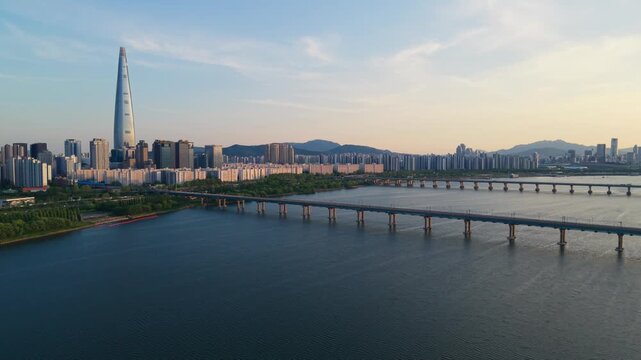 Aerial rise approaching Jamsil Railway Bridge and sunlit Lotte World Tower over the Han River at scenic golden sunset with Seoul skyline and mountains in South Korea