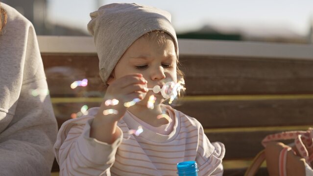 Little girl in a beanie blowing soap bubbles in warm daylight, playful and carefree outdoors moment capturing joyful childhood, concentration and innocent wonder