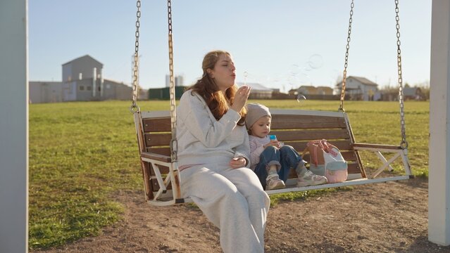 Mother and young daughter enjoying bonding time together on a swing outdoors, laughing while blowing soap bubbles and creating happy childhood memories in the sunshine