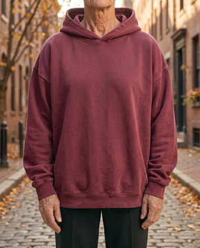 Outdoor portrait of man wearing blank maroon oversized hoodie mockup, commercial template on brick street with autumn trees, city background