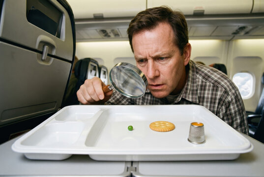 Sarcastic photo of passenger using magnifying glass to inspect tiny meal portion on tray in airplane cabin. Shrinkflation, tiny serving size and airline catering rip-off concept layout