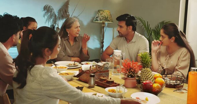 Indian happy relax joint family group sitting around table eat food talk speak share meal pick up glass drink water young adult male female mom girl boy child spend time enjoy night room home place