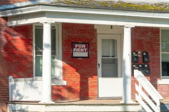 Historic red brick apartment building with a prominent 'FOR RENT' sign on its inviting shaded porch, featuring classic white columns, multiple mailboxes, and a moss-covered roof, perfect for real.