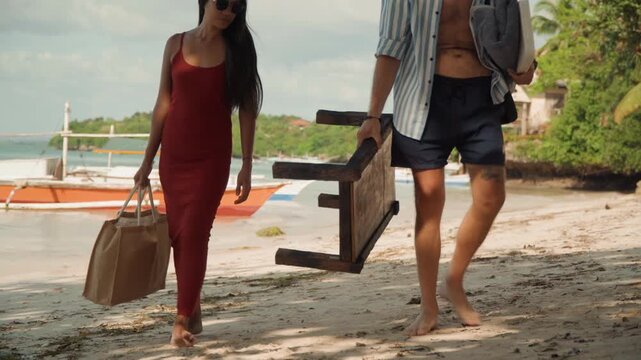 Couple walking on beach carrying chair, setting rustic wooden stool beside boat, woman in red dress holding straw bag, man in hat carrying folded chair, barefoot footprints in sand, palms and gentle