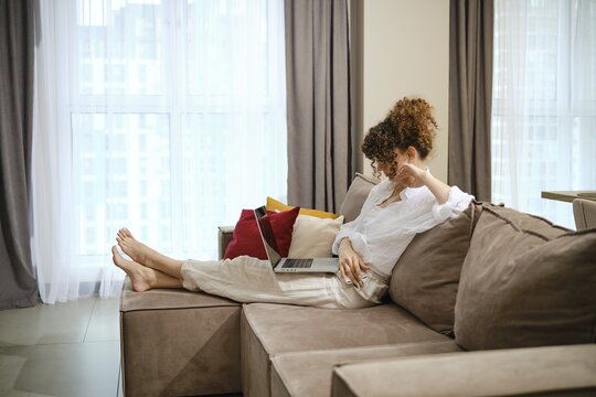 A young woman with curly hair is lounging on a stylish sofa in a bright living room and watching movie on a laptop. The atmosphere combines comfort with a modern design, creating a serene workspace at home