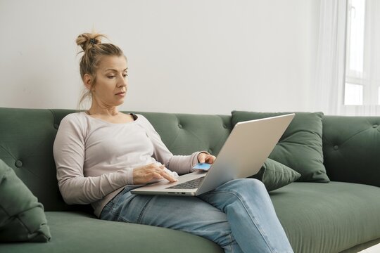 Middle-aged woman sitting on green couch with laptop and using credit card while shopping online at home
