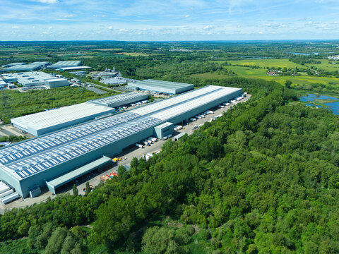 Aerial view of a large industrial warehouse and distribution center surrounded by lush green forest and fields in Coleshill, England, United Kingdom.